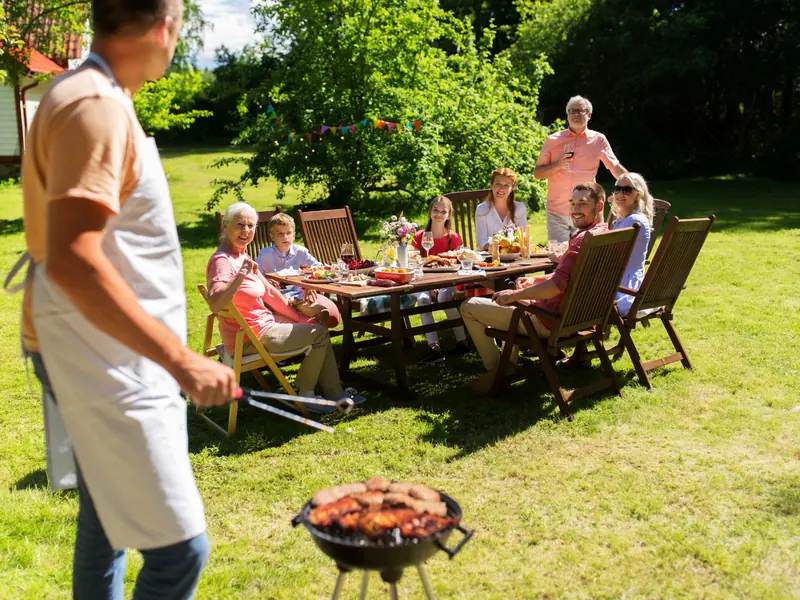 Familie an einer langen Tafel im Garten, im Vordergrund wird gegrillt
